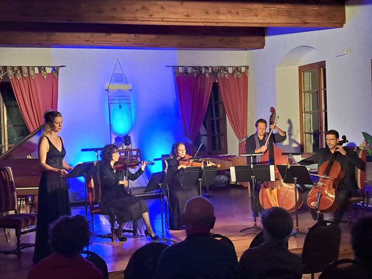 Violinists, a singer, and a cellist performing music inside the historic Priory, of Tufts European Center, in Talloires, surrounded by rustic stone walls and wooden beams.