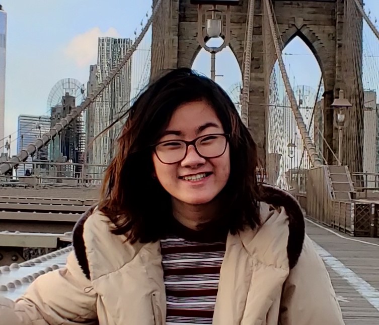 Trang, an Asian woman with black hair and glasses, smiles at the camera for a portrait with the Brooklyn Bridge behind her.